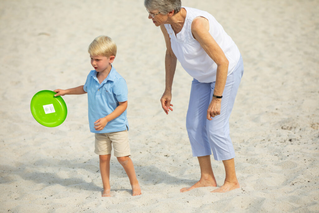 An elderly woman and a young boy stand barefoot on a sandy beach. The boy holds a green frisbee, and both appear to be playing together. The woman is slightly bent forward, watching the boy. Vero Beach Photoshoot