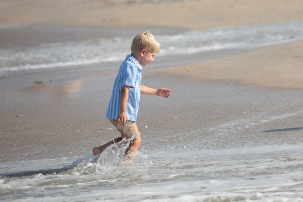 A young boy with blond hair wearing a blue striped shirt and tan shorts runs and splashes through shallow ocean waves on a sandy beach. Vero Beach Photoshoot