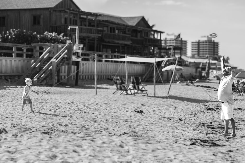 A child and an adult play with a frisbee on a sandy beach. Wooden buildings, empty chairs, and tall structures are visible in the background on a sunny day. Vero Beach Photoshoot