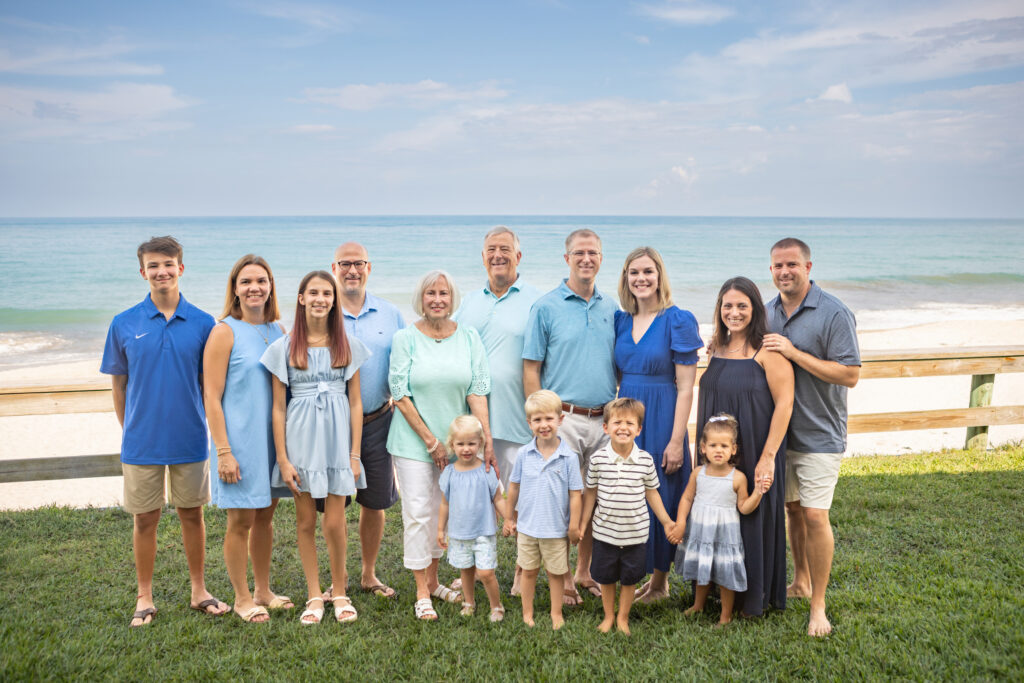 Thirteen people of various ages pose together on grass near a beach. They wear blue, white, and neutral-toned clothes. Ocean waves and a blue sky are in the background. Vero Beach Photoshoot