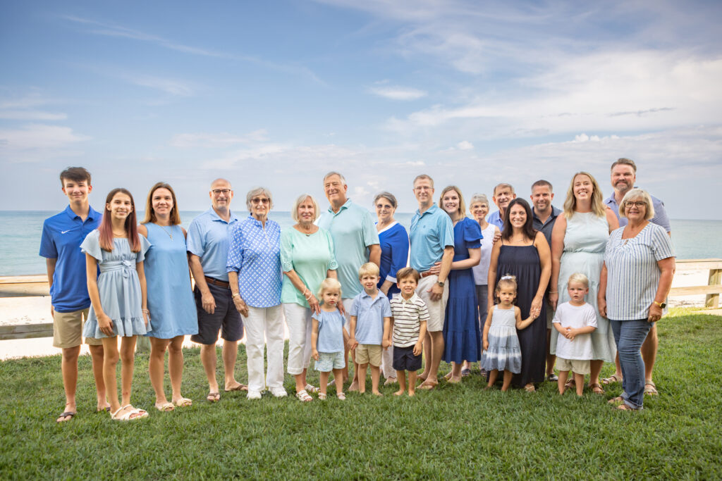 A large multigenerational family poses together outside on grass by the ocean, all smiling and dressed in coordinated blue and white outfits under a partly cloudy sky. Vero Beach Photoshoot