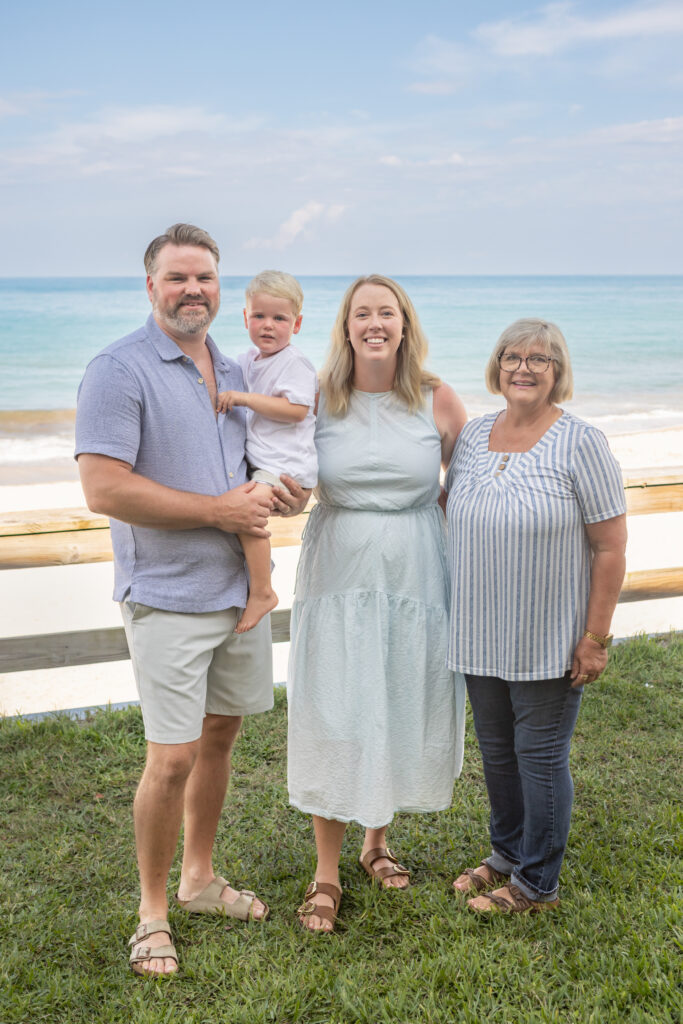 Four people stand on grass by a beach: a man holds a young child, with a woman in a light dress and an older woman in a striped shirt, all smiling in front of the ocean and sky. Vero Beach Photoshoot