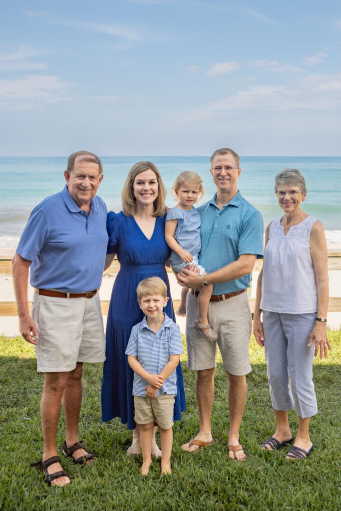 Three adults, two children, and an elderly woman stand smiling on grass by the beach, all dressed in blue and white summer clothes, with the ocean and sky in the background. Vero Beach Photoshoot