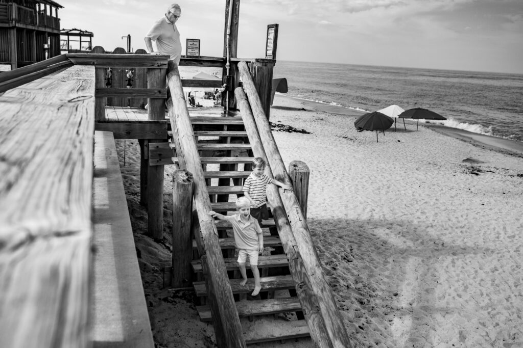 Two children descend wooden stairs to a sandy beach while an adult looks on from above. Umbrellas and the ocean are visible in the background under a cloudy sky. Vero Beach Photoshoot