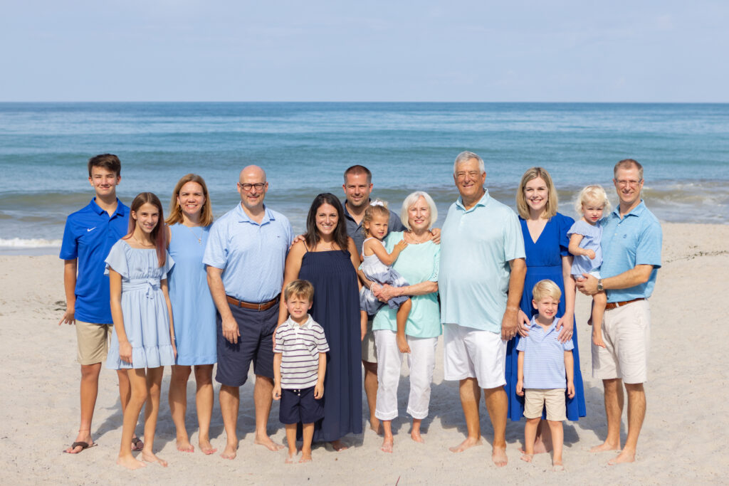 Fourteen people of various ages pose and smile on a sandy beach with the ocean in the background. Most wear blue, white, or light-colored clothing. It is a sunny day with a clear sky. Vero Beach Photoshoot